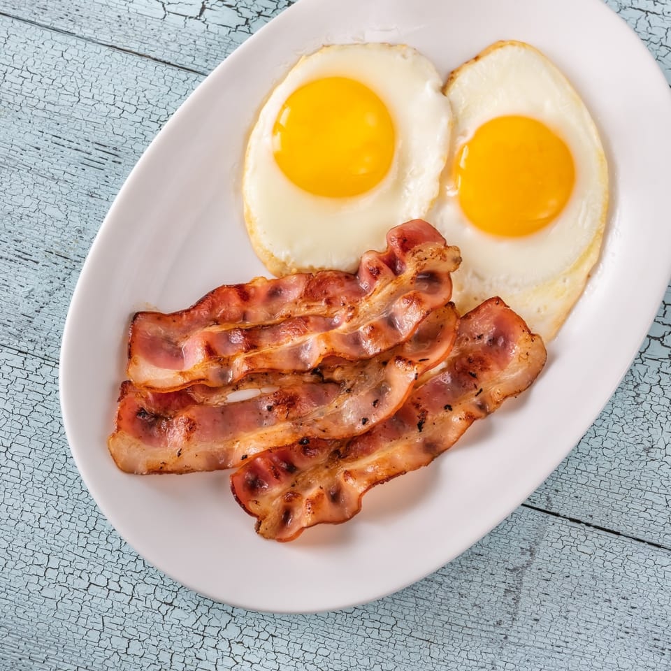 A white plate holds two sunny-side-up eggs and several strips of crispy bacon, a classic diner breakfast.