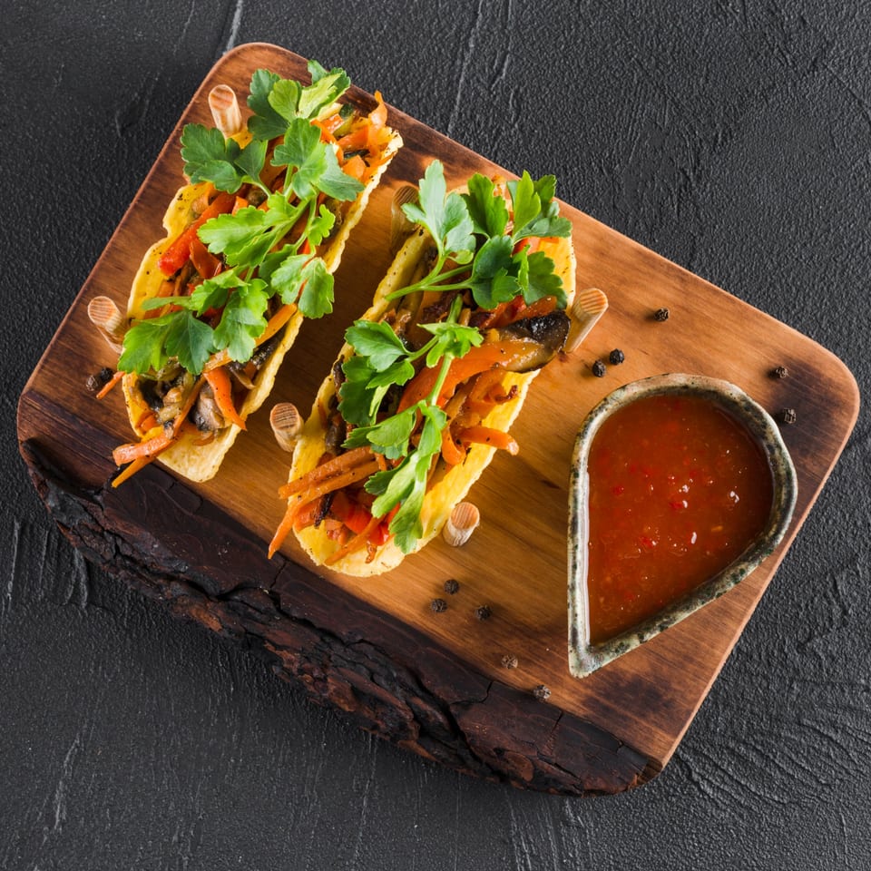 Two tacos filled with vegetables and parsley, served with a side of red sauce on a wooden board.